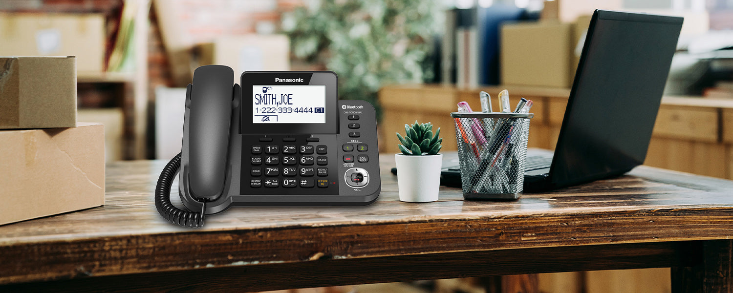 a corded desk phone sits on top of a wooden desk which includes an open laptop and pens in a penholder